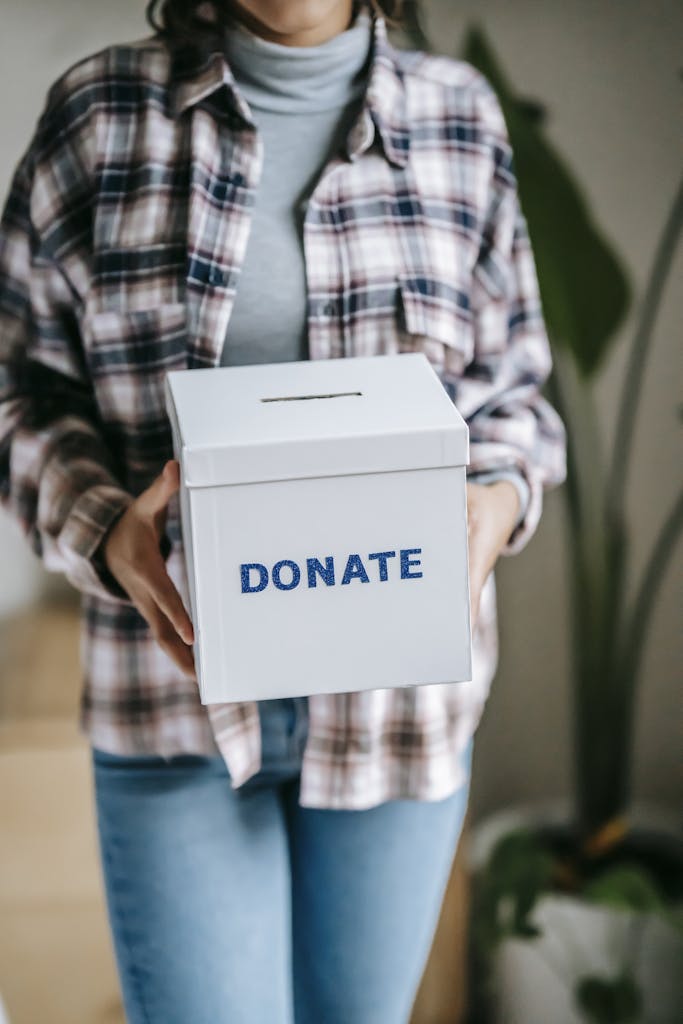 Crop unrecognizable woman in casual clothes standing with box for donations in hands on light room
